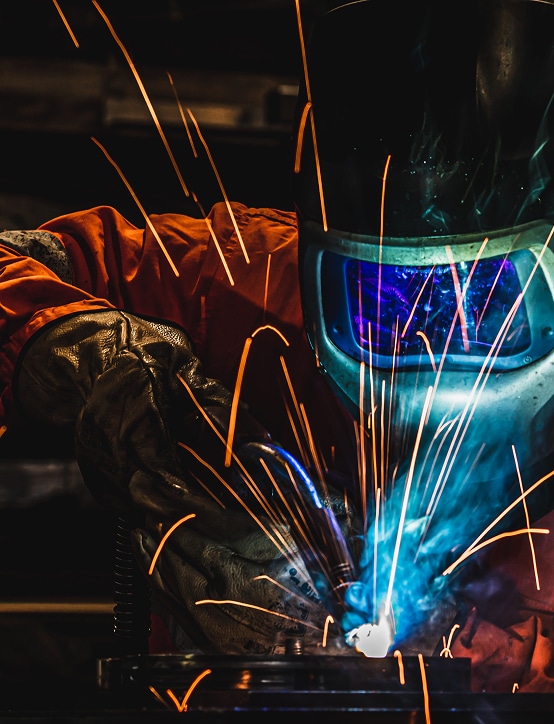 Soudeur en action : l'intensité de la soudure à l'arc. Soudeur en équipement orange et gants, réalisant une soudure. Étincelles vives et arc électrique bleu traversant le masque de protection.