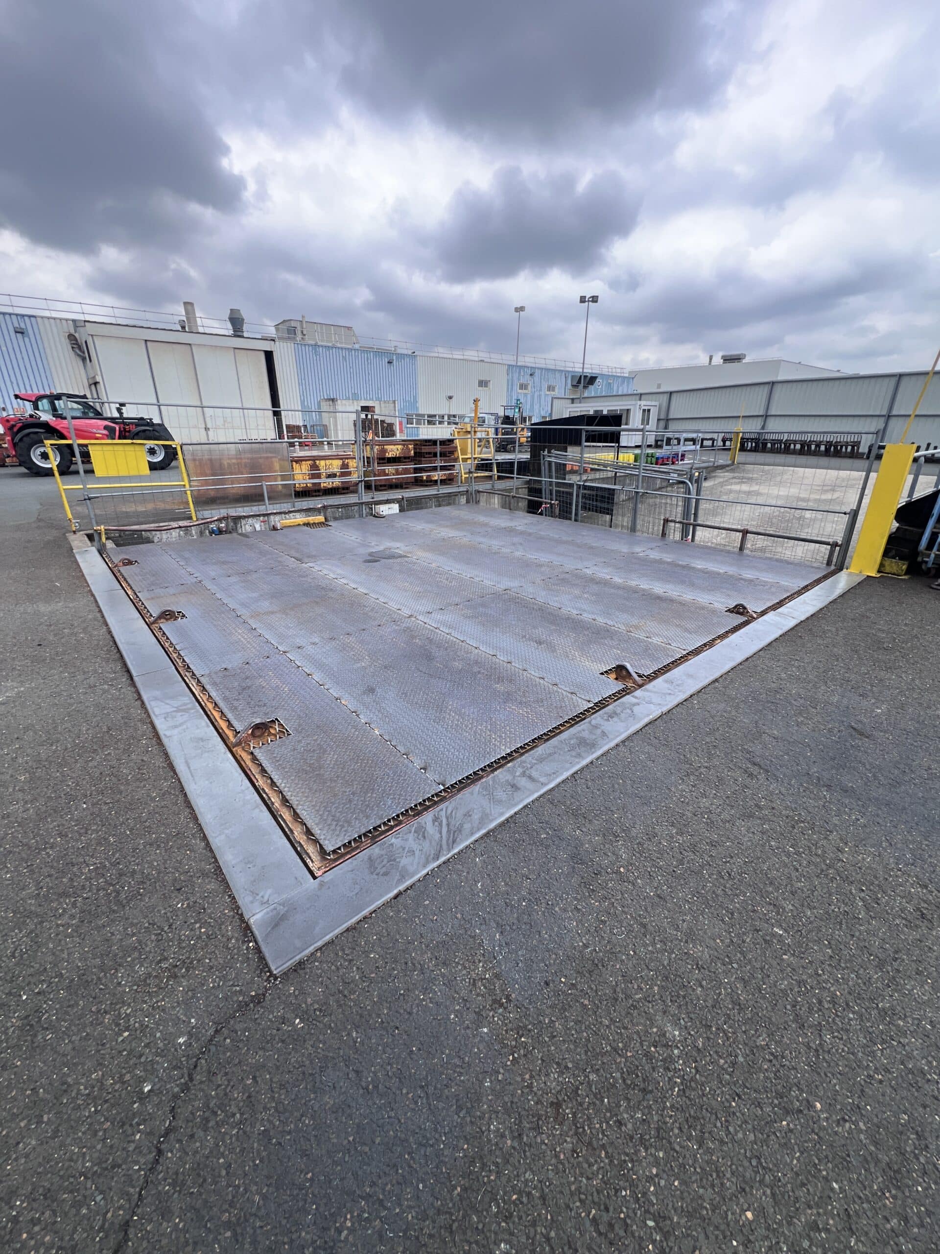 Plateforme de pesage industrielle en métal strié devant une usine avec un chariot télescopique rouge.