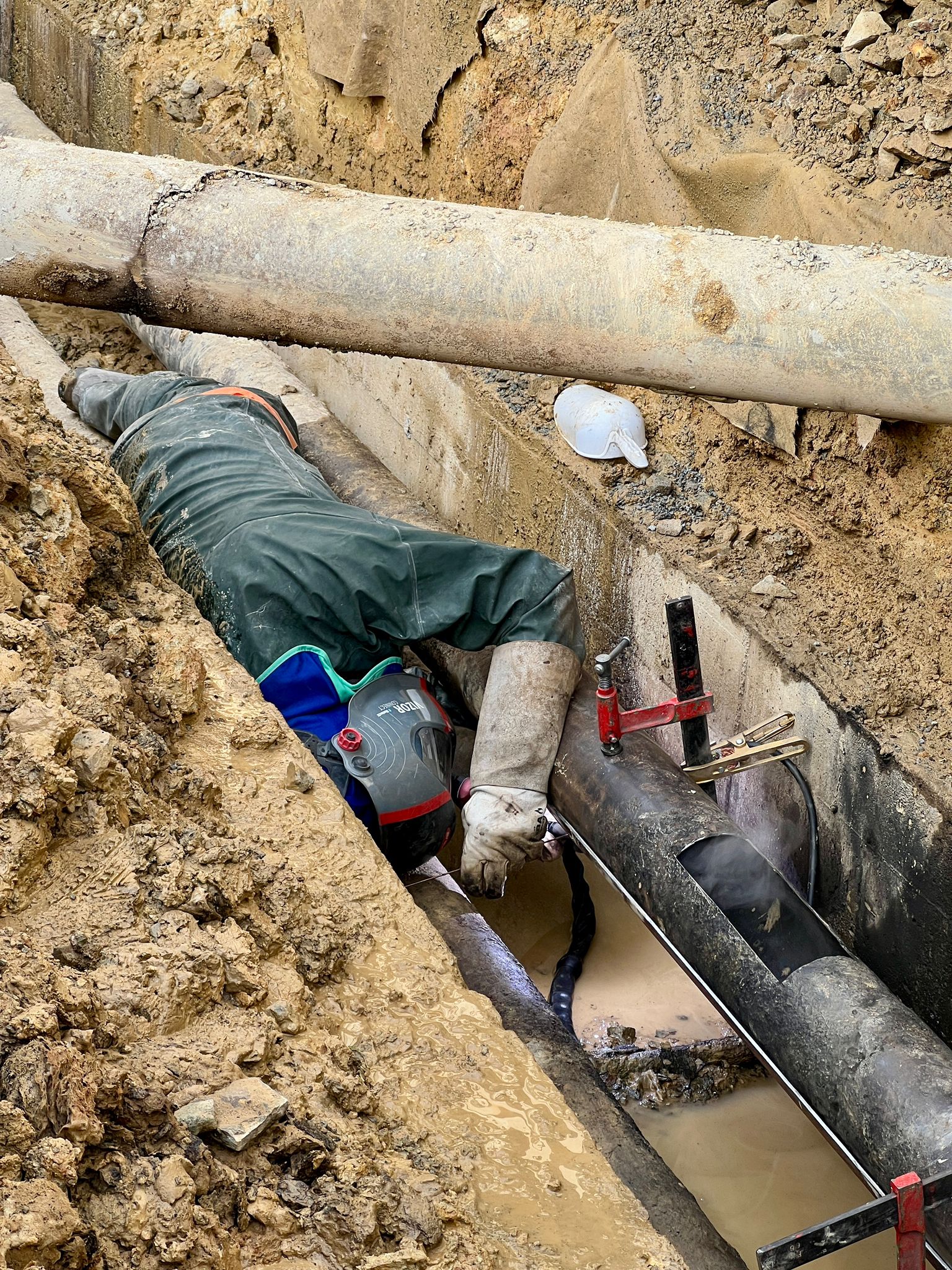 Soudeur équipé réparant une canalisation dans une tranchée étroite et boueuse lors de travaux de maintenance publique.